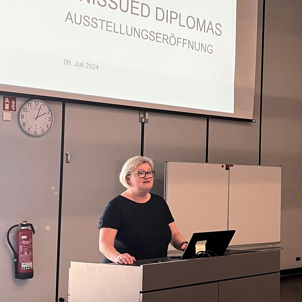 Prof. Dr. Eva Waller steht am Rednerpult. Hinter ihr steht auf einer Präsentationsfolie "Unissued Diplomas. Ausstellungseröffnung"