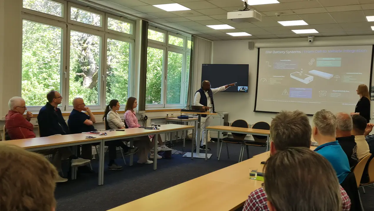 Eine Präsentation in einem Raum mit mehreren Tischen. Ein Referent zeigt auf eine Folie, die Informationen zu Batterien enthält. Das Publikum, bestehend aus Menschen unterschiedlichen Alters, hört aufmerksam zu und blickt in Richtung der Präsentation.