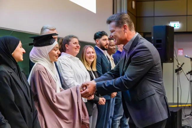 Eine Gruppe von Absolventen steht auf einer Bühne, während eine Absolventin in einem Abschlussoutfit einem Lehrenden die Hand schüttelt. Im Hintergrund stehen weitere Personen, die applaudieren oder lächeln. Die Szene vermittelt eine Feier des Abschlusses und der Erfolge.