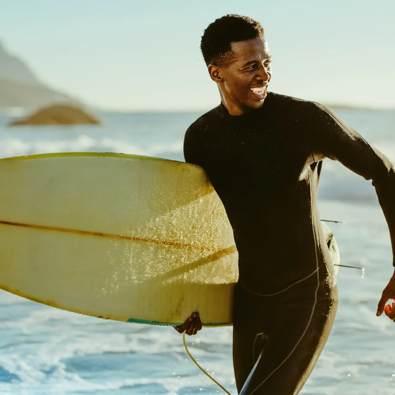 Ein Surfer in einem schwarzen Neoprenanzug hält ein Surfbrett und lächelt. Der Hintergrund zeigt das Meer mit Wellen und eine Küstenlandschaft. Das Licht ist warm und der Himmel klar, was eine entspannte Atmosphäre vermittelt.