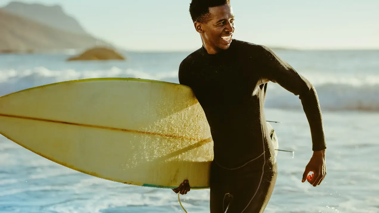 Ein Surfer in einem schwarzen Neoprenanzug hält ein Surfbrett und lächelt. Der Hintergrund zeigt das Meer mit Wellen und eine Küstenlandschaft. Das Licht ist warm und der Himmel klar, was eine entspannte Atmosphäre vermittelt.