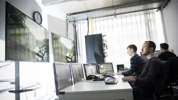 In einem modernen Büro sitzen zwei Männer an einem Tisch, vor mehreren Monitoren. Sie arbeiten konzentriert, während im Hintergrund natürliche Elemente sichtbar sind. An der Wand hängt eine Uhr, die die Tageszeit anzeigt. Licht fällt durch große Fenster, die den Raum erhellen.