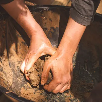 Two hands knead damp earth in a bowl. The hands are dirty as the person concentrates on shaping the material.