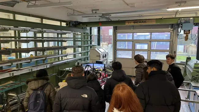Eine Gruppe von Personen in Winterjacken steht in einem technischen Raum um den Fahrsimulator des studentischen Rennteams Scuderia Mensa der Hochschule RheinMain. Sie beobachten ein Projekt, das im Zusammenhang mit Technik und Maschinenbau steht. Der Raum ist hell erleuchtet und gut ausgestattet.