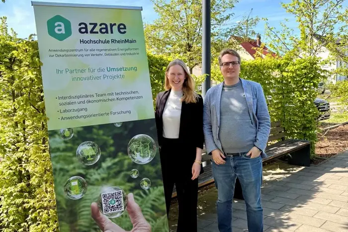 Zwei Personen stehen neben einem Banner der Hochschule RheinMain mit dem Schriftzug "azare". Das Banner bewirbt innovative Projekte und zeigt eine QR-Code. Im Hintergrund sind Bäume und blauer Himmel sichtbar.