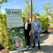 Zwei Personen stehen neben einem Banner der Hochschule RheinMain mit dem Schriftzug "azare". Das Banner bewirbt innovative Projekte und zeigt eine QR-Code. Im Hintergrund sind Bäume und blauer Himmel sichtbar.
