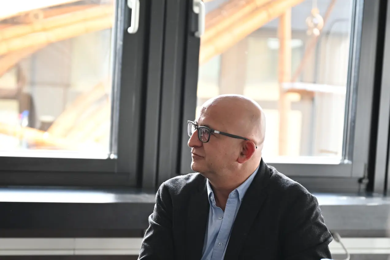Zwei Männer sitzen an einem Tisch und diskutieren. Der Mann links trägt einen Anzug und gestikuliert während des Sprechens, der Mann rechts hört aufmerksam zu. Auf dem Tisch stehen Getränke und eine Auswahl an Gebäck. Hinter den Männern sind große Fenster mit Tageslicht zu sehen.