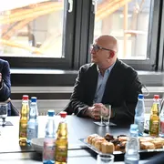 Zwei Männer sitzen an einem Tisch und diskutieren. Der Mann links trägt einen Anzug und gestikuliert während des Sprechens, der Mann rechts hört aufmerksam zu. Auf dem Tisch stehen Getränke und eine Auswahl an Gebäck. Hinter den Männern sind große Fenster mit Tageslicht zu sehen.