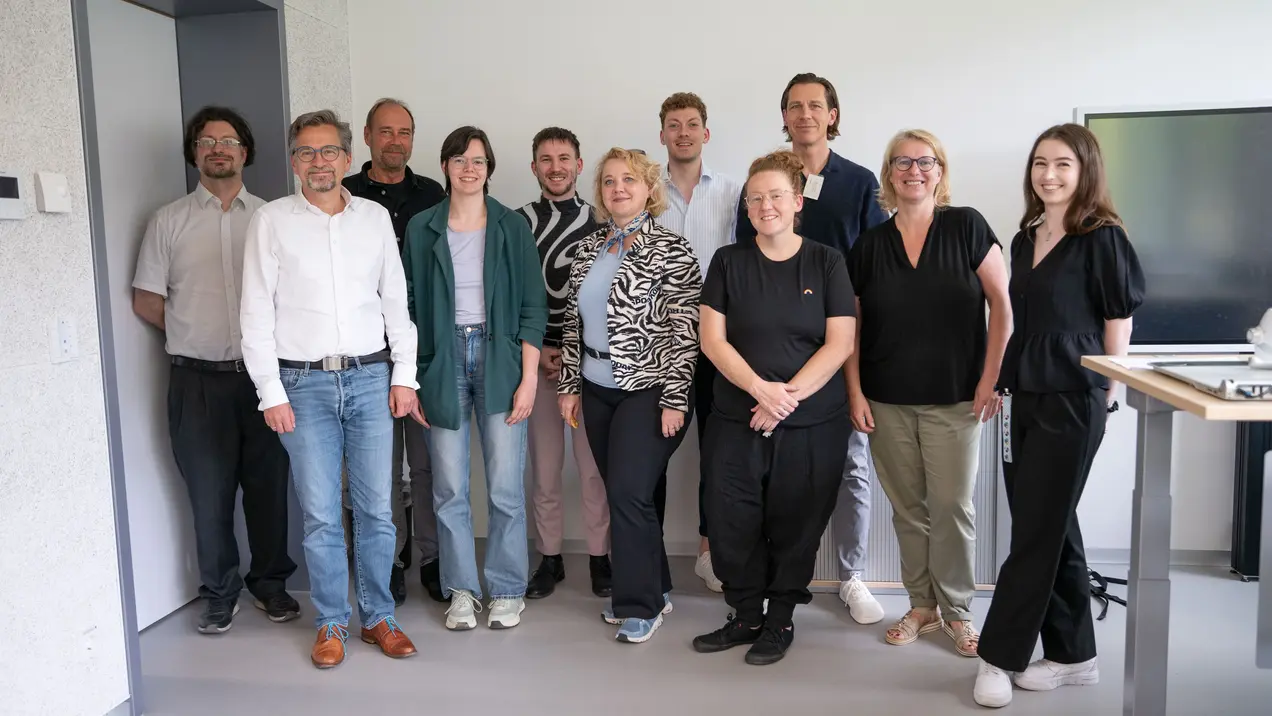 Eine Gruppe von zwölf Personen steht in einem modernen Raum. Die Menschen zeigen unterschiedliche Ausdrucksformen, einige lächeln. Sie tragen legere Kleidung. Im Hintergrund sind einige Büromöbel und ein Bildschirm zu sehen.