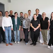 Eine Gruppe von zwölf Personen steht in einem modernen Raum. Die Menschen zeigen unterschiedliche Ausdrucksformen, einige lächeln. Sie tragen legere Kleidung. Im Hintergrund sind einige Büromöbel und ein Bildschirm zu sehen.