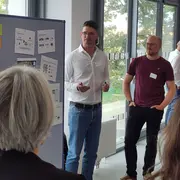 Eine Gruppe von vier Personen steht in einem modernen Raum. Ein Mann in einem weißen Hemd präsentiert vor einem aufgestellten Poster. Die anderen Zuhörer, welche teilweise mit dem Rücken zur Kamera stehen, hören aufmerksam zu. Das Poster enthält verschiedene Informationen und Grafiken.
