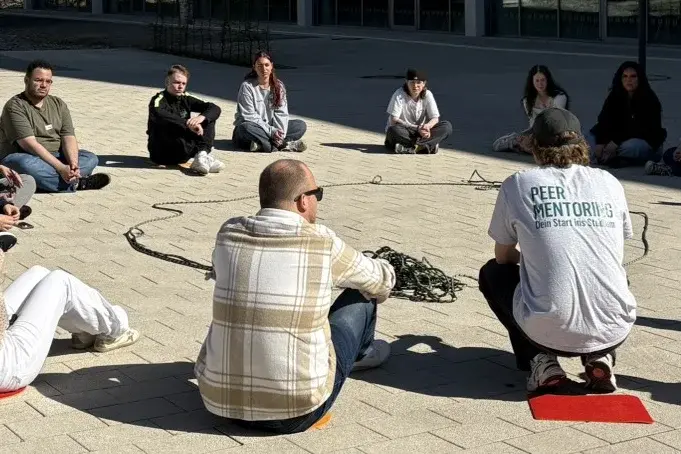 Eine Gruppe von 15 Personen sitzt in einem Halbkreis auf dem Boden vor einem modernen Gebäude. Sie scheinen an einer Diskussion oder Aktivität im Freien beteiligt zu sein. Der Himmel ist klar und blau.