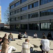 Eine Gruppe von 15 Personen sitzt in einem Halbkreis auf dem Boden vor einem modernen Gebäude. Sie scheinen an einer Diskussion oder Aktivität im Freien beteiligt zu sein. Der Himmel ist klar und blau.