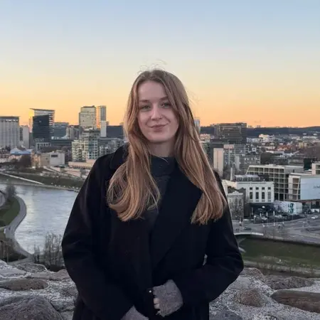 Eine Frau mit langen, lockigen Haaren steht in einem schwarzen Mantel auf einer Steinmauer. Im Hintergrund erstreckt sich eine Stadtlandschaft mit einem Fluss und einem klaren Himmel bei Sonnenuntergang.