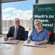 Zwei Personen sitzen an einem Tisch und unterschreiben Dokumente. Im Hintergrund ist ein Banner der Hochschule RheinMain mit dem Slogan „Mach’s zu deinem Business!“ sichtbar. Das Bild vermittelt eine Atmosphäre der Zusammenarbeit und des Formalitäts.