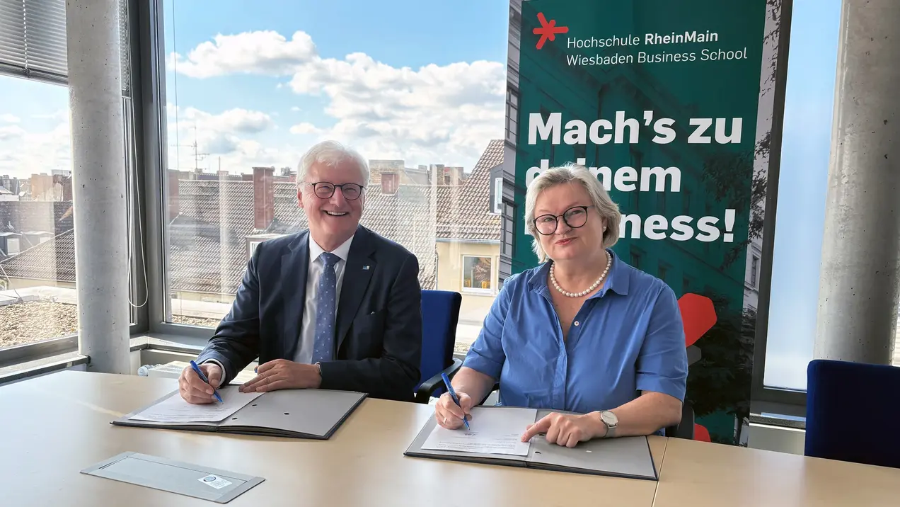 Zwei Personen sitzen an einem Tisch und unterschreiben Dokumente. Im Hintergrund ist ein Banner der Hochschule RheinMain mit dem Slogan „Mach’s zu deinem Business!“ sichtbar. Das Bild vermittelt eine Atmosphäre der Zusammenarbeit und des Formalitäts.