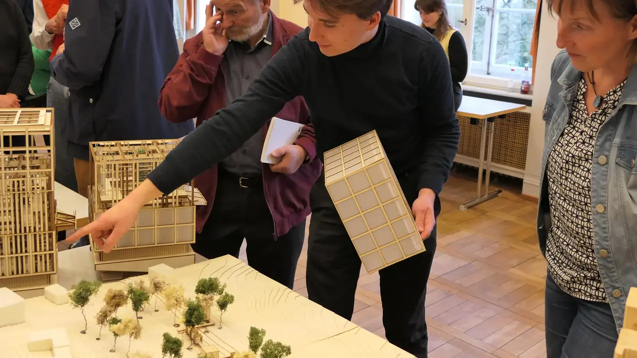 A group of people are looking at models of buildings on a table. A man is pointing at a model while a woman and another man listen attentively. Trees and more models can be seen in the background.