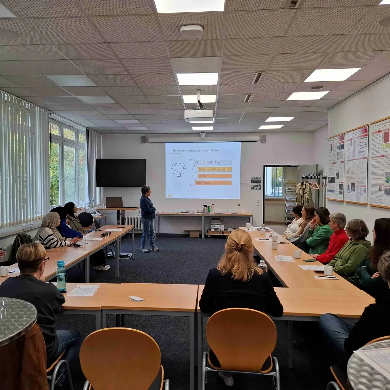 Ein Vortrag in einem Seminarraum, in dem eine Person vor einer Gruppe von Teilnehmern steht. Die Präsentation zeigt ein Diagramm an der Wand. Die Zuschauer sind in Reihen angeordnet und hören aufmerksam zu.