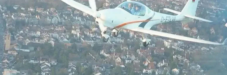 Ein kleines Flugzeug fliegt über eine städtische Landschaft. Im Cockpit sind zwei Piloten sichtbar. Die Umgebung zeigt eine Ansammlung von Gebäuden und Bäumen, die aus der Vogelperspektive zu sehen sind.