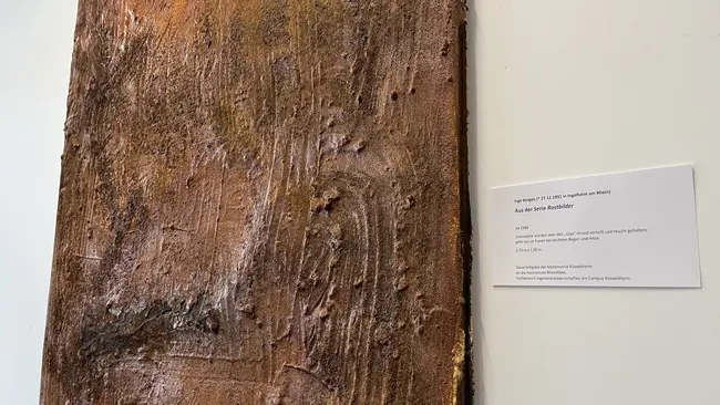 Ein rechtwinkliges, strukturiertes Wandbild aus rostbraunem Material, das texturierte Oberflächen und Linien aufweist. Es hängt an einer weißen Wand, daneben befindet sich ein Beschilderungsschild.