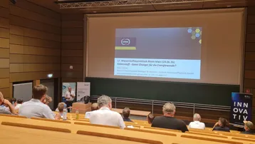 In einem Sitzungssaal findet eine Präsentation zum Thema "Trends und Innovationen" statt. Im Vordergrund sind mehrere Personen zu sehen, die dem Referenten auf der Bühne zuhören. Eine Präsentation zeigt den Titel und das Datum der Veranstaltung an der Wand.