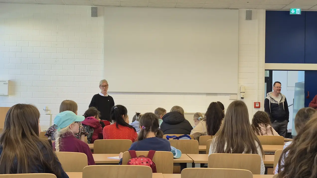 Im Vordergrund stehen zwei Personen, die vor einer Gruppe von Zuhörern sprechen. Die Zuhörer sind in einem Klassenraum mit Tischen und Stühlen angeordnet und richten ihre Aufmerksamkeit auf die Redner. Der Raum hat hohe Fenster und eine helle, einladende Atmosphäre.