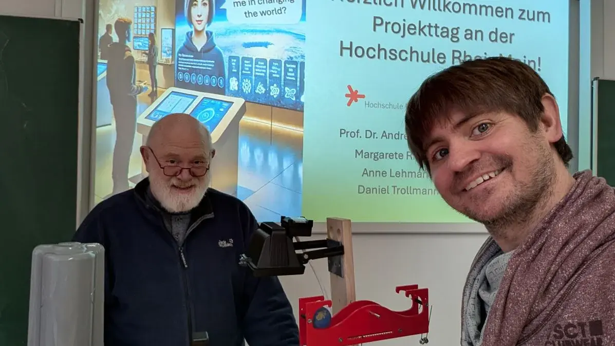 Ein älterer Mann mit Brille und ein jüngerer Mann stehen vor einem Bildschirm, der Grüße zum Projekt-Tag an der Hochschule Bielefeld anzeigt. Beide lächeln in die Kamera, während im Hintergrund verschiedene technische Geräte zu sehen sind.