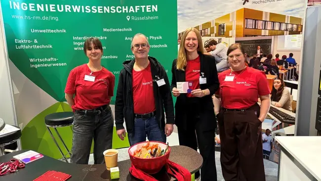 Auf der Messe stehen vier Personen am Stand der Hochschule RheinMain. Sie tragen rote T-Shirts und präsentieren Informationen über Ingenieurwissenschaften. Im Hintergrund sind Banner und eine moderne Gebäudeansicht zu sehen. Es liegt eine Schüssel mit bunten Stiften auf einem Tisch.