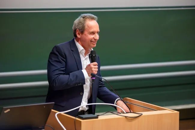 Ein lächelnder Mann steht an einem Rednerpult und hält ein Mikrofon in der Hand. Er trägt einen dunkelblauen Anzug und spricht zu einem Publikum. Im Hintergrund ist eine grüne Tafel sichtbar.