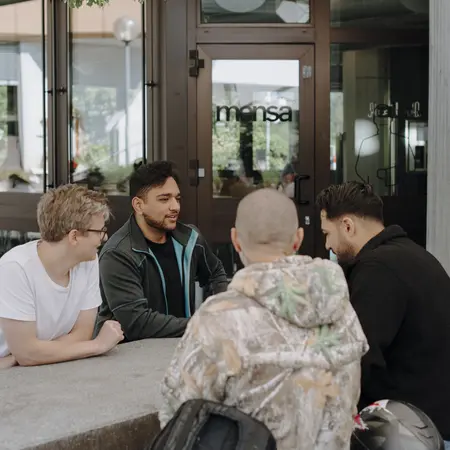 Fünf junge Männer sitzen an einem Tisch im Freien. Sie unterhalten sich lebhaft und wirken entspannt. Der Hintergrund zeigt große Fenster und Pflanzen, was eine angenehme Atmosphäre schafft.