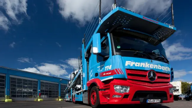 Ein blauer Mercedes-Lkw mit der Aufschrift "Frankenbach" steht vor einem Gebäude. Der Lkw hat eine spezielle Konstruktion für den Transport. Im Hintergrund sind Wolken und das Gebäude sichtbar.