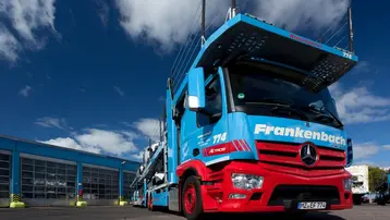 Ein blauer Mercedes-Lkw mit der Aufschrift "Frankenbach" steht vor einem Gebäude. Der Lkw hat eine spezielle Konstruktion für den Transport. Im Hintergrund sind Wolken und das Gebäude sichtbar.
