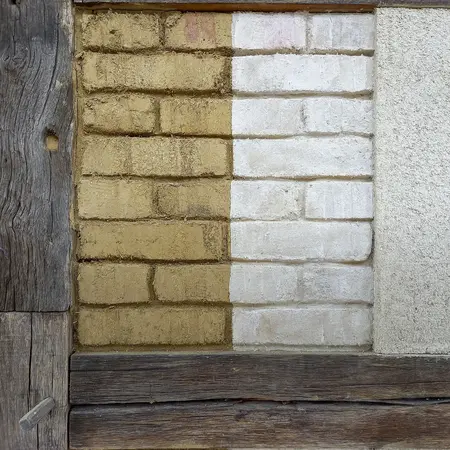 Eine Wand zeigt vier verschiedene Bauoberflächen: Links ein brauner Lehmputz, daneben ein weißer Ziegel, gefolgt von einem beige Putz und schließlich einer grauen Fläche. Die Textur und Farbe der Materialien variieren, um unterschiedlichen Bauweisen darzustellen. Es handelt sich um den Wandaufbau eines Fachwerk-Musterhaus im Hessenpark.
