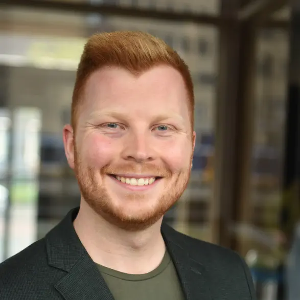Portrait von Mobilitätsmangement-Alumni Tim Kirschbaum. Er trägt einen schwarzen Blazer über einem dunkelgrünen T-Shirt. Im Hintergrund sind unscharfe Gebäude und Fenster zu erkennen.