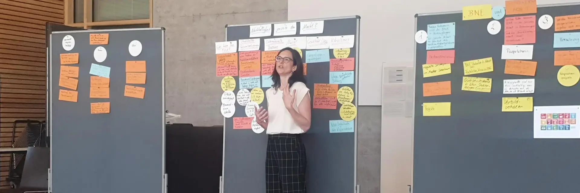 Eine Frau steht vor drei Stellwänden, die mit bunten Haftnotizen und Informationen beklebt sind. Sie hält eine Notiz in der Hand und erklärt vermutlich den Inhalt der Wände. Der Raum hat einen modernen, hellen Eindruck.