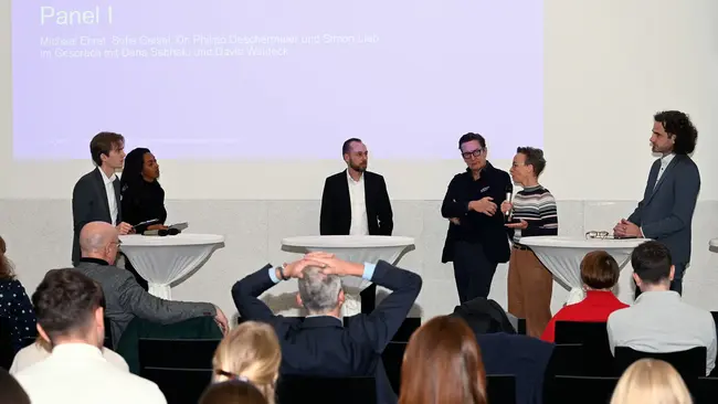 Ein Panel mit Michael Ehret, Sofie Geisel, Dr. Philipp Deschermeier, Simon Lieb, Dena Sebhatu und David Waldeck steht an Stehtischen beim 2. Wiesbadener Tag für sozial nachhaltiges Investieren an der Hochschule RheinMain. Sie diskutieren zu einem Thema, während das Publikum darauf achtet. 