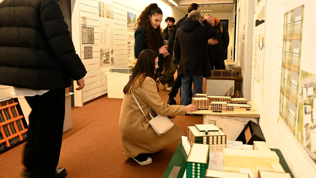 Architekturstudierende und weitere Interessierte besichtigen eine Ausstellung über die Architekturprojekte der Studierenden der HSRM. Eine Frau in einem beigen Mantel sitzt und betrachtet Modellbauten, während andere Besucher in der Nähe stehen und sich unterhalten. Die Wände sind mit Architekturzeichnungen und Plänen dekoriert.