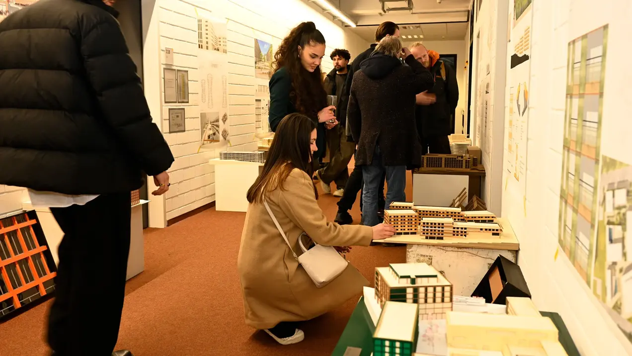Architecture students and other interested parties visit an exhibition on the architectural projects of HSRM students. A woman in a beige coat sits and looks at model buildings, while other visitors stand nearby and chat. The walls are decorated with architectural drawings and plans.