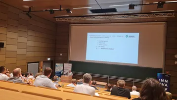 Eine Präsentation in einem Seminarraum. Auf der Leinwand sind Themen wie „Entwicklung“ und „Ergebnisse“ abgebildet, die mit dem Begriff „azare“ verbunden sind. Im Vordergrund sitzen zahlreiche Zuhörer und richten ihre Aufmerksamkeit auf den Referenten.