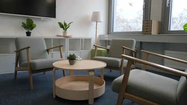 Der Raum zeigt eine entspannte Sitzecke mit zwei hellgrünen Sesseln und einem runden Tisch in der Mitte. Auf dem Tisch steht eine kleine Pflanze. Die Wände sind hell und ein Fenster bietet Blick nach draußen, mit natürlichem Licht, das den Raum erhellt.