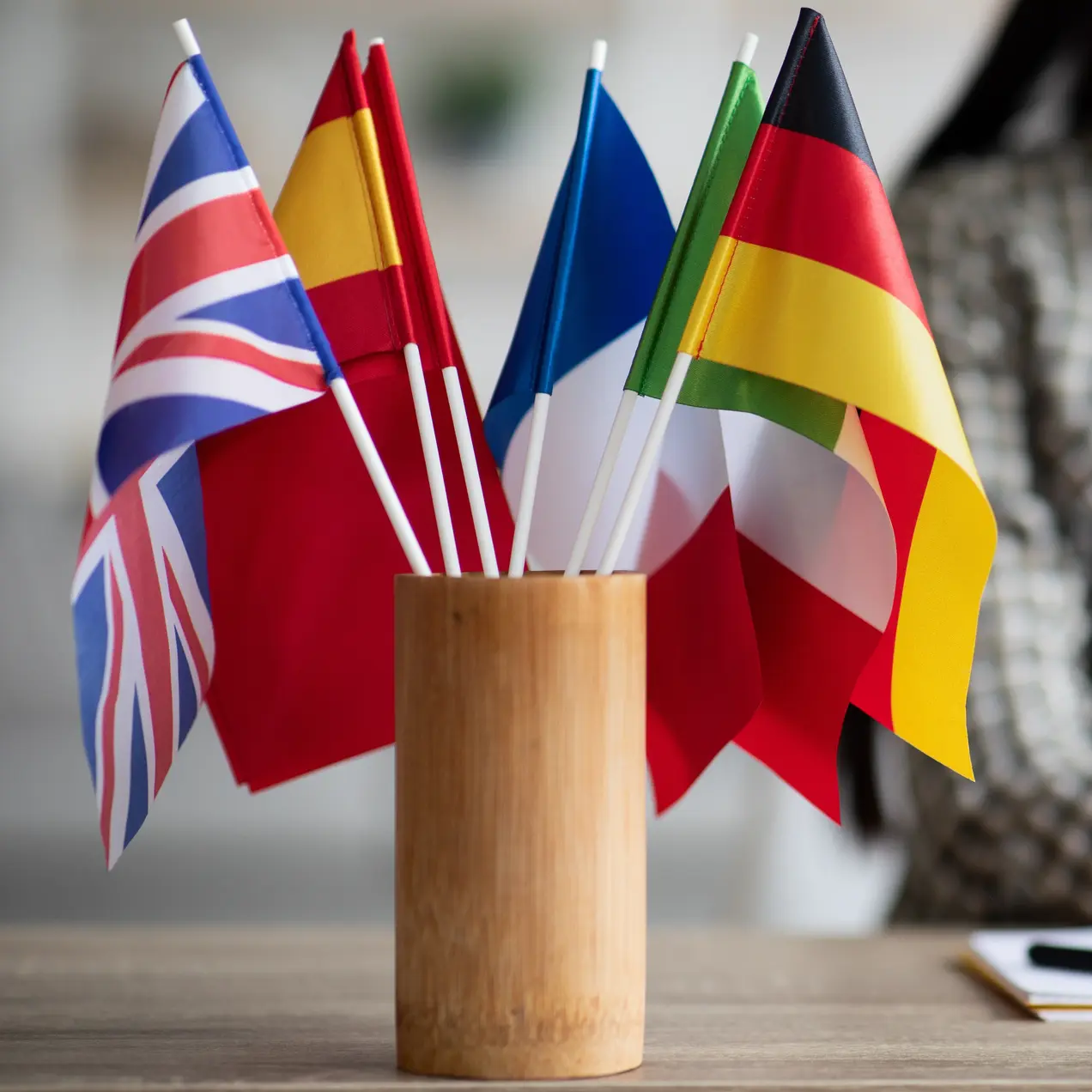 Auf dem Tisch steht ein Holzständer mit mehreren kleinen Flaggen, darunter die Flaggen von Großbritannien, Spanien, Frankreich, Italien und Deutschland. Im Hintergrund arbeitet eine Frau an einem Laptop.