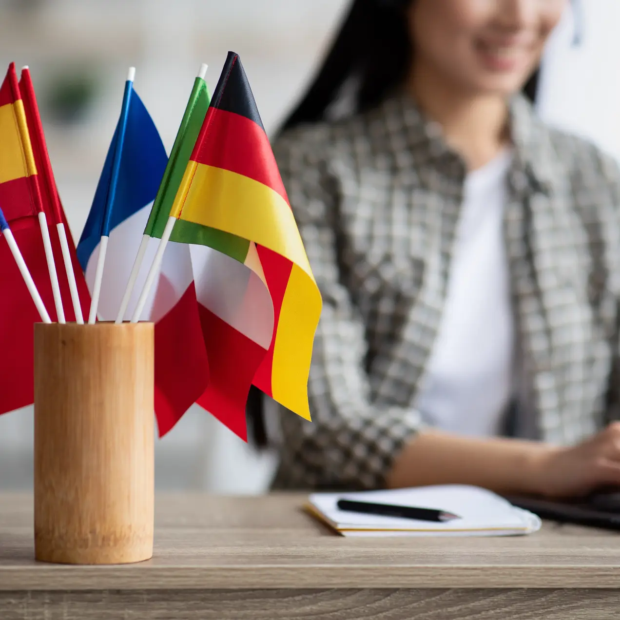 Auf dem Tisch steht ein Holzständer mit mehreren kleinen Flaggen, darunter die Flaggen von Großbritannien, Spanien, Frankreich, Italien und Deutschland. Im Hintergrund arbeitet eine Frau an einem Laptop.