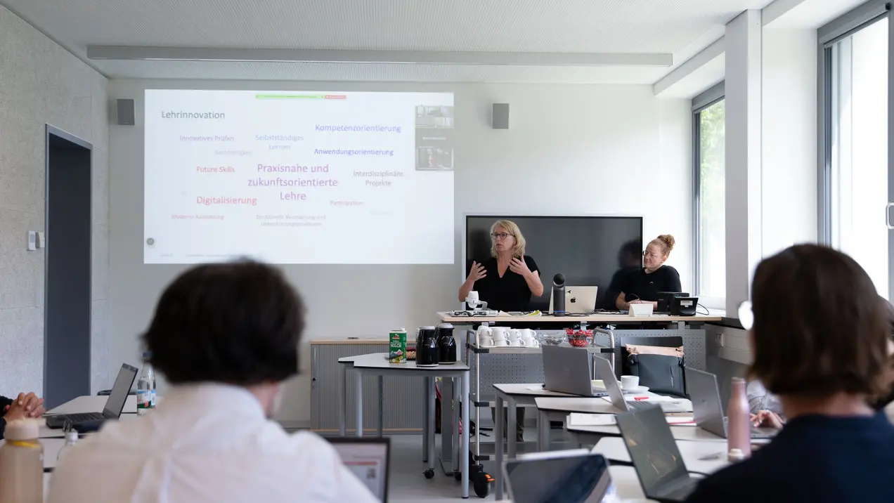 Eine Präsentation in einem modernen Schulungsraum. Eine Frau steht vorne und präsentiert, während eine zweite Person am Tisch sitzt. Auf einer Projektionsfläche sind Schlüsselbegriffe zu den Themen Lebensinnovationen und digitale Lehre zu sehen. Die Teilnehmenden arbeiten an Laptops.