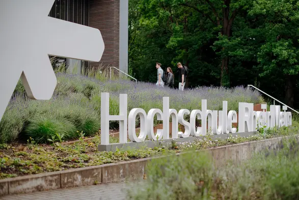 Ein Schild mit der Aufschrift „Hochschule RheinMain“ im Vordergrund auf dem Campus am Kurt-Schumacher-Ring. Im Hintergrund sind Studierende zu sehen, die einen Weg entlanggehen, der zur Hochschule führt.