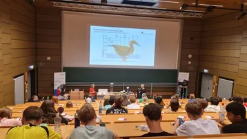 In einem Vortragssaal sitzen zahlreiche Zuhörer. Auf der Leinwand wird eine Grafik gezeigt, die einen Vogel und statistische Daten darstellt. Vor der Leinwand stehen zwei Referenten, die das Thema präsentieren.