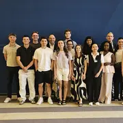 Gruppenfoto von 18 Personen, die zusammen stehen. Sie lächeln und tragen verschiedene Sommerkleidung. Im Hintergrund ist das Logo des Bundesministeriums der Finanzen sichtbar. Die Atmosphäre wirkt freundlich und einladend.