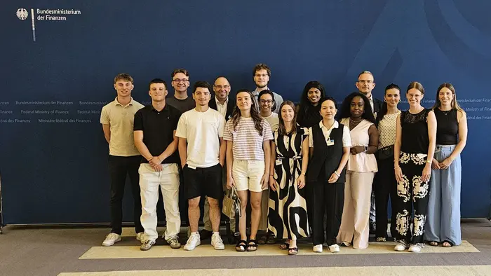 Gruppenfoto von 18 Personen, die zusammen stehen. Sie lächeln und tragen verschiedene Sommerkleidung. Im Hintergrund ist das Logo des Bundesministeriums der Finanzen sichtbar. Die Atmosphäre wirkt freundlich und einladend.