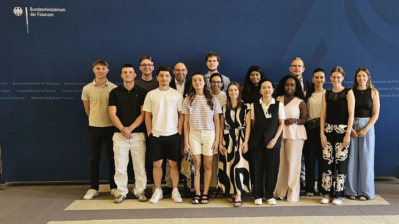 Gruppenfoto von 18 Personen, die zusammen stehen. Sie lächeln und tragen verschiedene Sommerkleidung. Im Hintergrund ist das Logo des Bundesministeriums der Finanzen sichtbar. Die Atmosphäre wirkt freundlich und einladend.