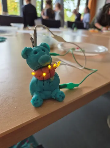 Eine kleine, aus Knetmasse geformte Bärfigur in Türkis steht auf einem Tisch. Sie hat leuchtende, orangefarbene Augen und trägt einen roten Ball im Mund. Vom Körper der Figur führen Drähte mit LED-Leuchten zu einer Stromquelle. Im Hintergrund sind unscharfe Personen zu sehen.