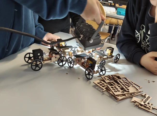 Eine Gruppe von Personen arbeitet an einem Roboterfahrzeug aus Holz. Eine Hand hält ein Werkzeug, während andere Hände Teile halten und zusammenbauen. Um das Fahrzeug liegen weitere Holzplatten mit vormontierten Teilen.